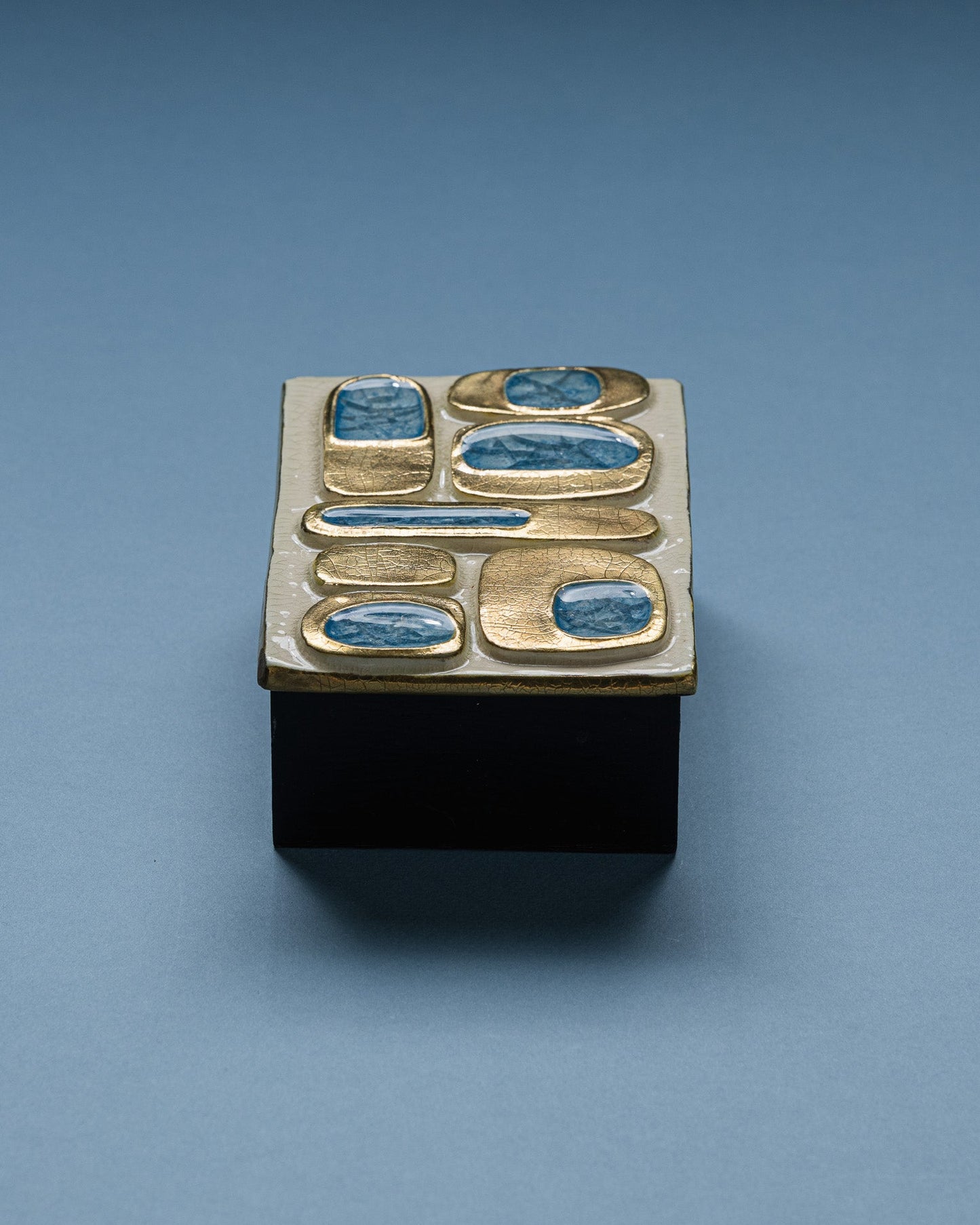Boîte en céramique émaillée de Mithé Espelt réalisée dans les années 1960. Couvercle décoré de cabochons en verre bleu incrustés dans des alvéoles dorées à l'émail craquelé, sur fond ivoire. Base en bois peint noir.
Très bel état.