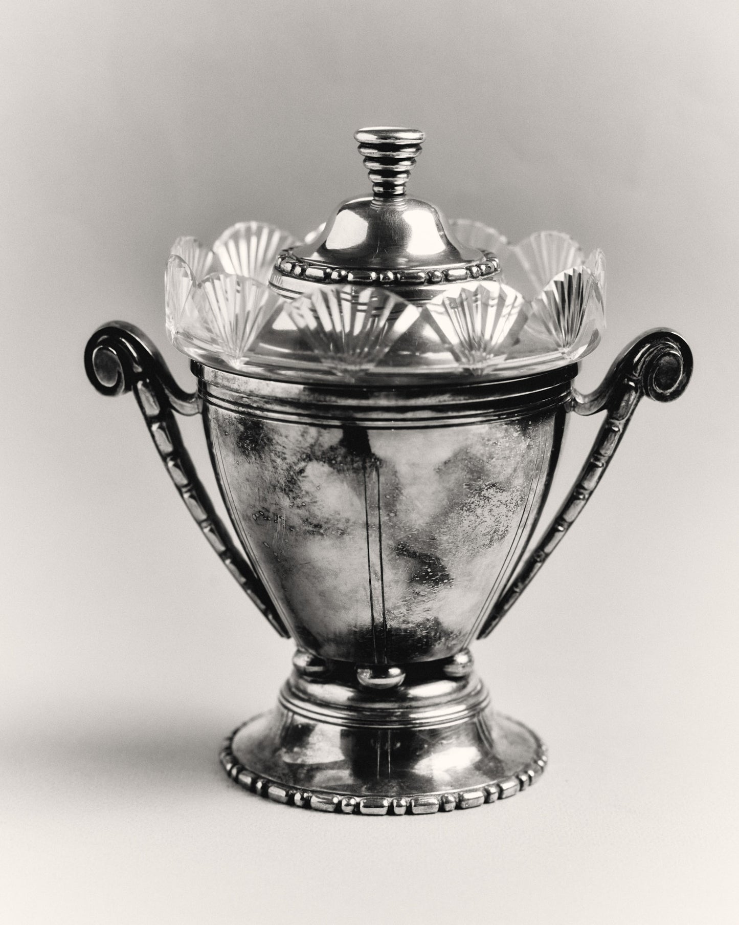 Bonbonnière ou sucrier à couvercle en métal argenté et verre taillé. Le corps en verre est bordé d'un col à pans rayonnants. Monture et couvercle en métal argenté à frises parlées, anses latérales en volute. Poinçonné.
Bon état général, légère patine d'usage.