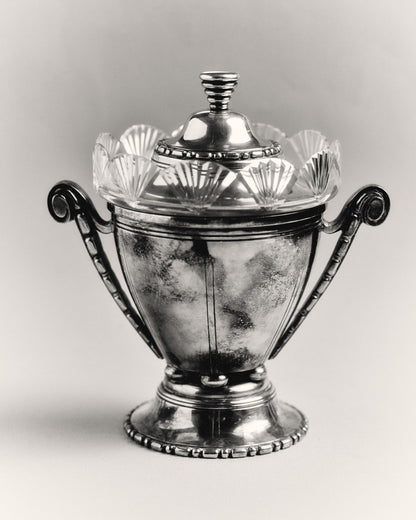 Bonbonnière ou sucrier à couvercle en métal argenté et verre taillé. Le corps en verre est bordé d'un col à pans rayonnants. Monture et couvercle en métal argenté à frises parlées, anses latérales en volute. Poinçonné.
Bon état général, légère patine d'usage.