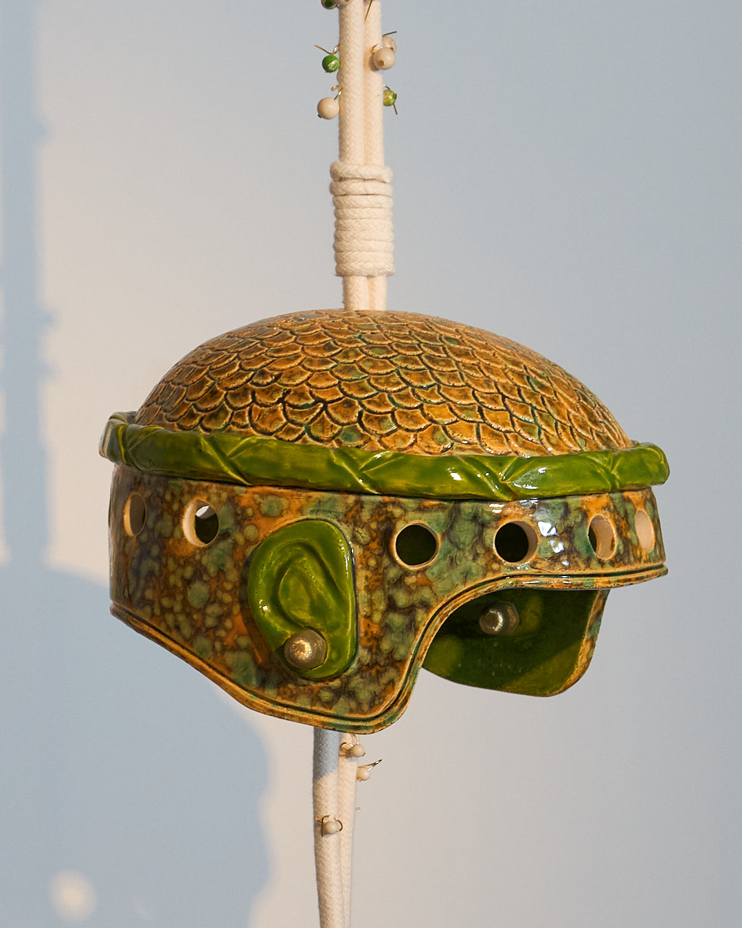 Casque en céramique émaillée, inspiré d’un casque militaire où cheveux et oreilles se dessinent sous la matière, comme si la céramique avait capturé l’empreinte vivante de sa propriétaire. L’œuvre évoque à la fois puissance et délicatesse, un équilibre entre protection et ouverture.
Suspendue à une corde parsemée de perles semi-précieuses — jaspe fossile et régalite, pierres de protection et de guidance —, cette création associe la céramique émaillée à des éléments en laiton évoquant des boucles d’oreilles 