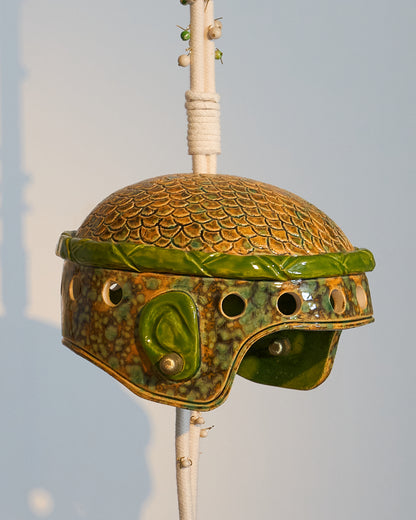 Casque en céramique émaillée, inspiré d’un casque militaire où cheveux et oreilles se dessinent sous la matière, comme si la céramique avait capturé l’empreinte vivante de sa propriétaire. L’œuvre évoque à la fois puissance et délicatesse, un équilibre entre protection et ouverture.
Suspendue à une corde parsemée de perles semi-précieuses — jaspe fossile et régalite, pierres de protection et de guidance —, cette création associe la céramique émaillée à des éléments en laiton évoquant des boucles d’oreilles 