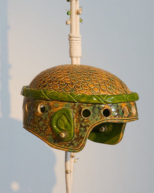 Casque en céramique émaillée, inspiré d’un casque militaire où cheveux et oreilles se dessinent sous la matière, comme si la céramique avait capturé l’empreinte vivante de sa propriétaire. L’œuvre évoque à la fois puissance et délicatesse, un équilibre entre protection et ouverture.
Suspendue à une corde parsemée de perles semi-précieuses — jaspe fossile et régalite, pierres de protection et de guidance —, cette création associe la céramique émaillée à des éléments en laiton évoquant des boucles d’oreilles 
