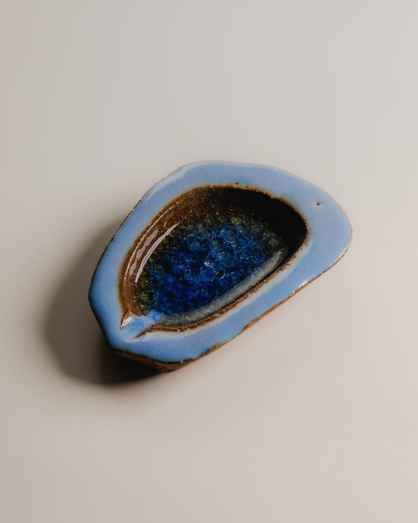 Cendrier de forme libre en céramique émaillée bleue, décoré en son centre d'une inclusion de verre fondu aux nuances de bleu profond et de brun ambré, formant un effet de craquelure et de transparence rappelant une géode. Travail artisanal des années 1970. Très bel état.