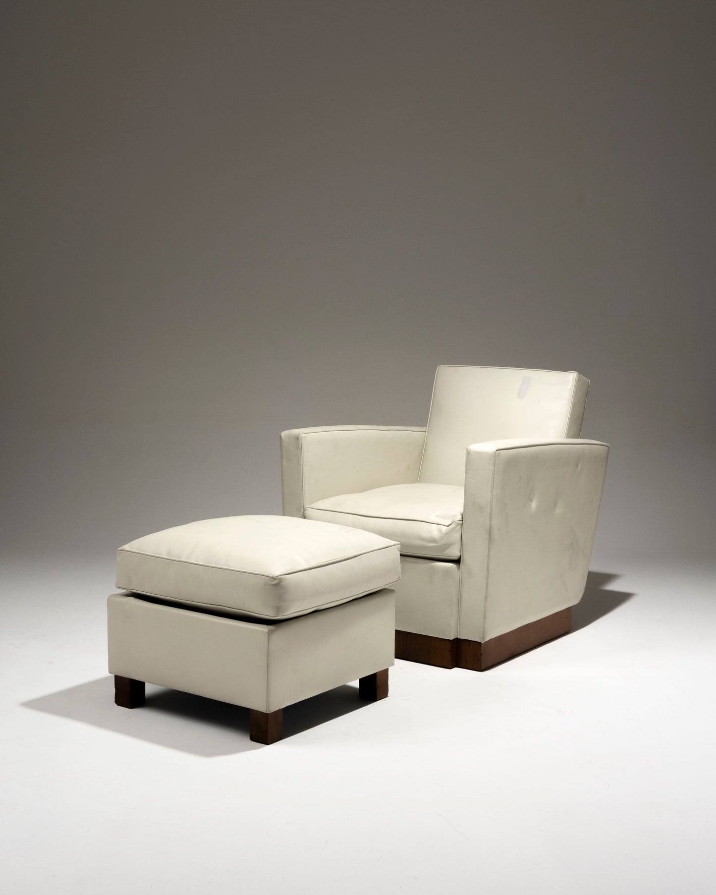 Fauteuil art déco et son repose-pied recouverts d'un cuir blanc cassé, par l'architecte et designer français Jacques Adnet (1900-1984) dans les années 1930. Le dossier incliné est encadré par deux accotoirs. Le piètement plein est en plaquage de noyer. Le repose pied est recouvert d'un coussin en plume. En bon état général malgré quelques altérations du cuir liées à l'usage et au temps.