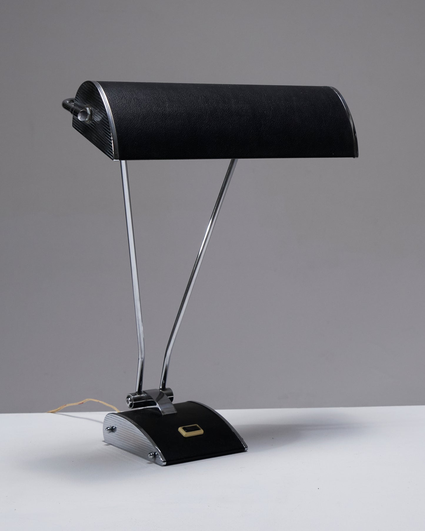 Lampe de bureau en tôle d'aluminium, conçue par Eileen Gray pour Jumo dans les années 1950. Son pied est équipé d'un interrupteur, et l'abat-jour effet cuir est réglable avec plusieurs inclinaisons possibles, offrant une grande flexibilité d'éclairage. Fonctionnelle, elle présente une combinaison de matériaux élégants et un design pratique. Prise européenne.