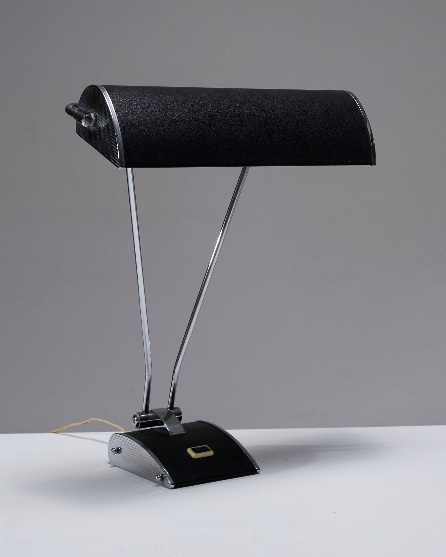 Lampe de bureau en tôle d'aluminium, conçue par Eileen Gray pour Jumo dans les années 1950. Son pied est équipé d'un interrupteur, et l'abat-jour effet cuir est réglable avec plusieurs inclinaisons possibles, offrant une grande flexibilité d'éclairage. Fonctionnelle, elle présente une combinaison de matériaux élégants et un design pratique. Prise européenne.