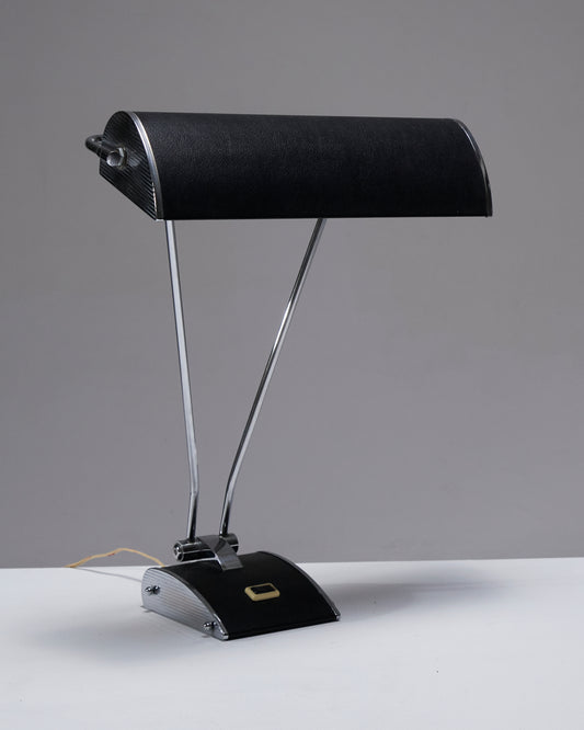 Lampe de bureau en tôle d'aluminium, conçue par Eileen Gray pour Jumo dans les années 1950. Son pied est équipé d'un interrupteur, et l'abat-jour effet cuir est réglable avec plusieurs inclinaisons possibles, offrant une grande flexibilité d'éclairage. Fonctionnelle, elle présente une combinaison de matériaux élégants et un design pratique. Prise européenne.