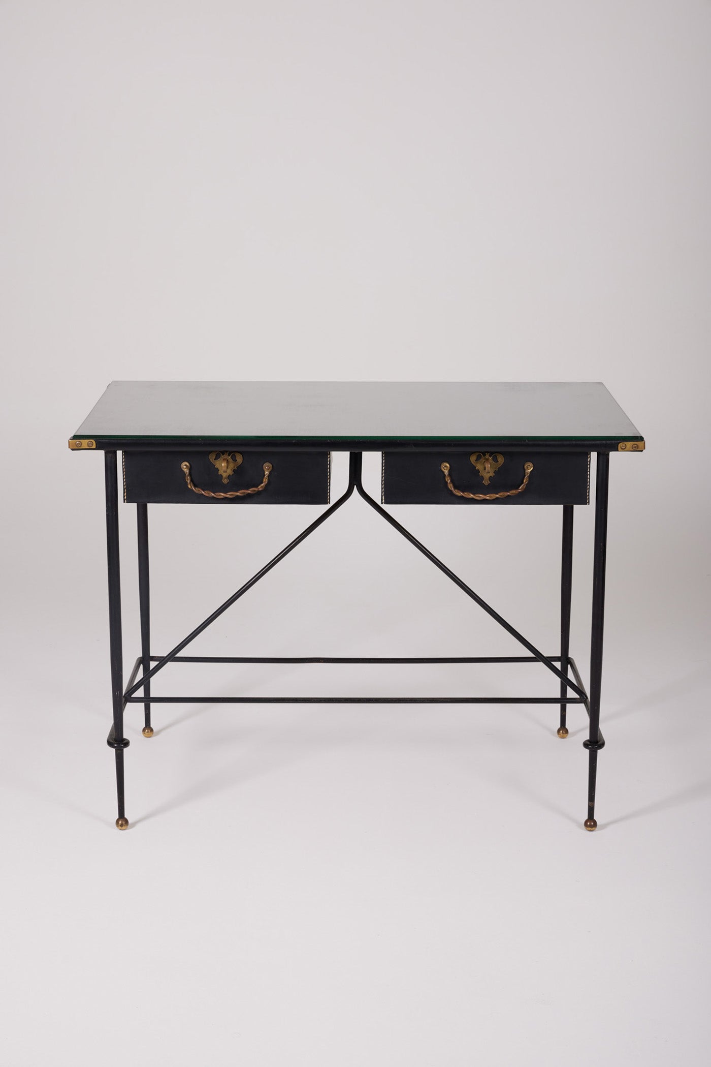  Bureau d'exception du designer Jacques Adnet (1900-1984), années 1940. Le piétement est en fer forgé patiné noir, le plateau en verre et les détails du bureau sont en cuir et laiton. Les deux tiroirs coulissants sont recouverts de cuir piqué à la main. Jacques Adnet a laissé une empreinte indélébile dans le monde du design avec son style distinctif et son innovation constante, il collabora notamment avec la maison Hermès sur des meubles en cuir de haute qualité, sa marque de fabrique.