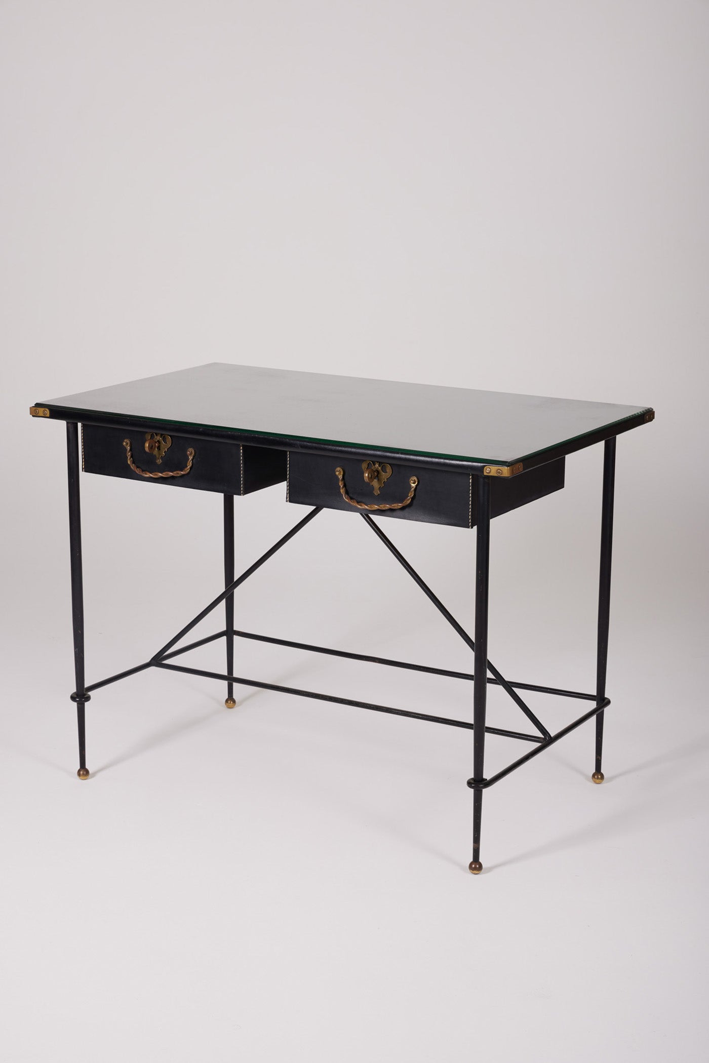  Bureau d'exception du designer Jacques Adnet (1900-1984), années 1940. Le piétement est en fer forgé patiné noir, le plateau en verre et les détails du bureau sont en cuir et laiton. Les deux tiroirs coulissants sont recouverts de cuir piqué à la main. Jacques Adnet a laissé une empreinte indélébile dans le monde du design avec son style distinctif et son innovation constante, il collabora notamment avec la maison Hermès sur des meubles en cuir de haute qualité, sa marque de fabrique.