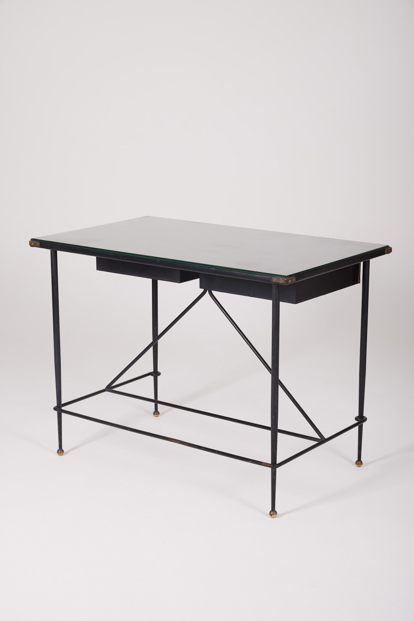  Bureau d'exception du designer Jacques Adnet (1900-1984), années 1940. Le piétement est en fer forgé patiné noir, le plateau en verre et les détails du bureau sont en cuir et laiton. Les deux tiroirs coulissants sont recouverts de cuir piqué à la main. Jacques Adnet a laissé une empreinte indélébile dans le monde du design avec son style distinctif et son innovation constante, il collabora notamment avec la maison Hermès sur des meubles en cuir de haute qualité, sa marque de fabrique.