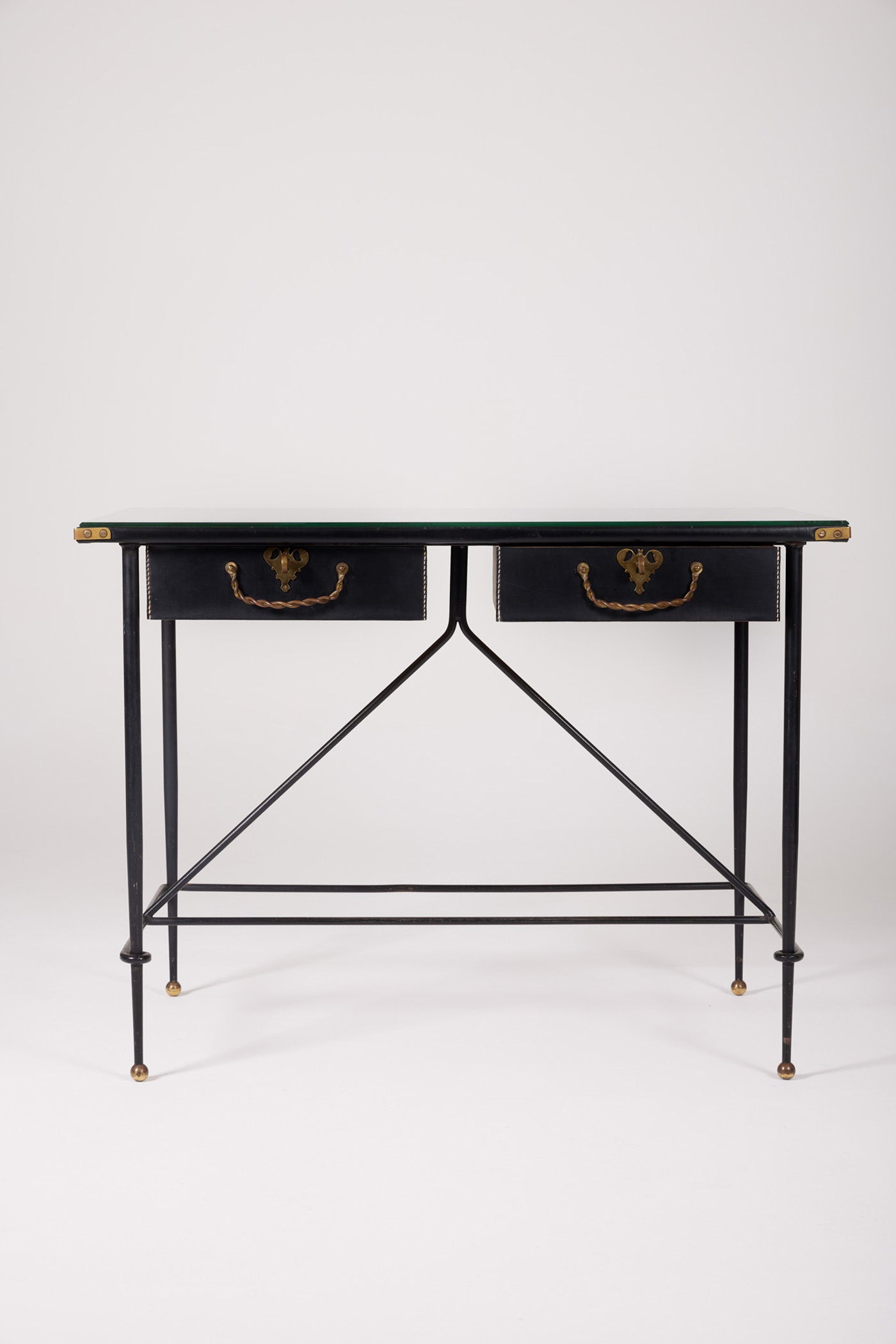  Bureau d'exception du designer Jacques Adnet (1900-1984), années 1940. Le piétement est en fer forgé patiné noir, le plateau en verre et les détails du bureau sont en cuir et laiton. Les deux tiroirs coulissants sont recouverts de cuir piqué à la main. Jacques Adnet a laissé une empreinte indélébile dans le monde du design avec son style distinctif et son innovation constante, il collabora notamment avec la maison Hermès sur des meubles en cuir de haute qualité, sa marque de fabrique.