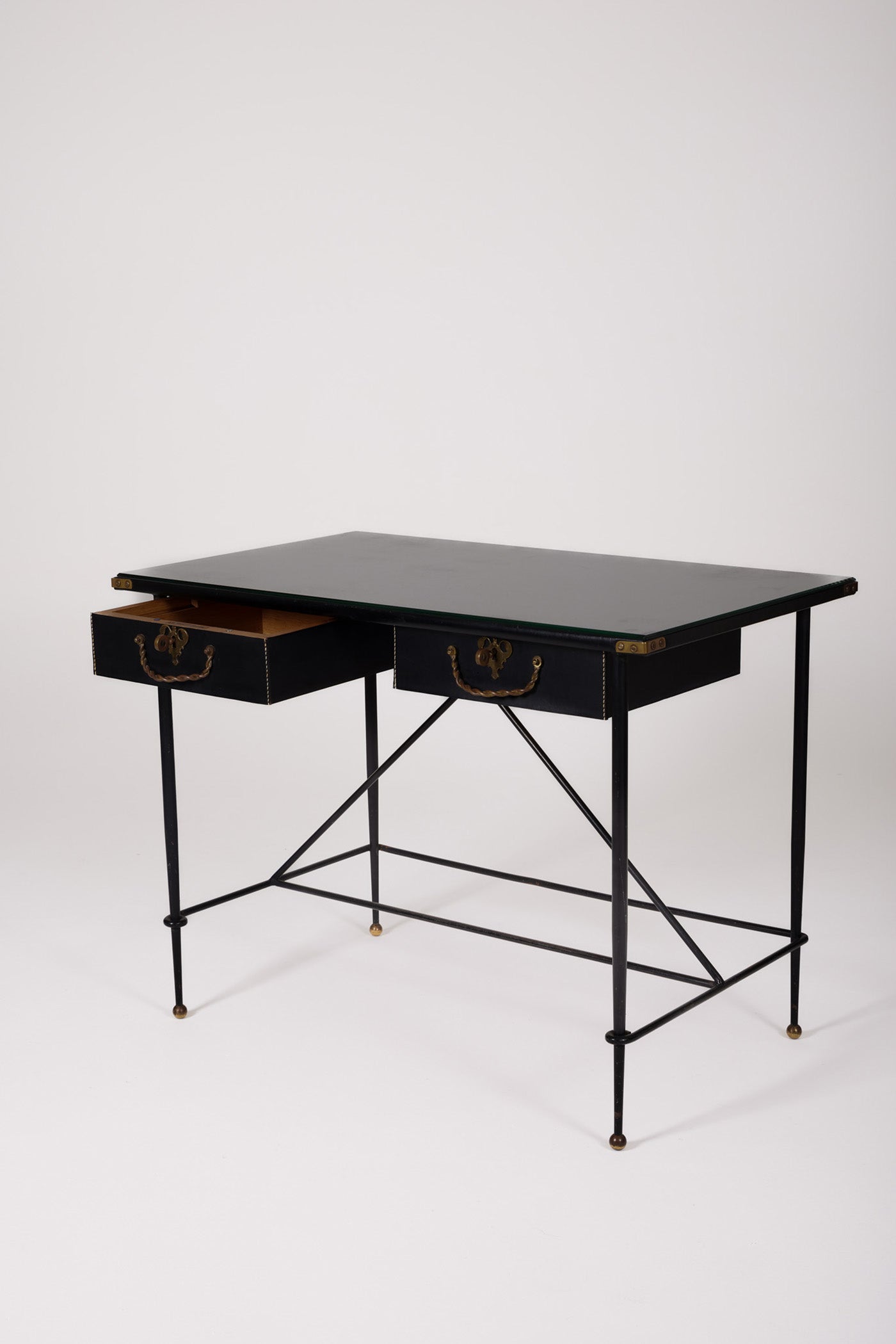  Bureau d'exception du designer Jacques Adnet (1900-1984), années 1940. Le piétement est en fer forgé patiné noir, le plateau en verre et les détails du bureau sont en cuir et laiton. Les deux tiroirs coulissants sont recouverts de cuir piqué à la main. Jacques Adnet a laissé une empreinte indélébile dans le monde du design avec son style distinctif et son innovation constante, il collabora notamment avec la maison Hermès sur des meubles en cuir de haute qualité, sa marque de fabrique.