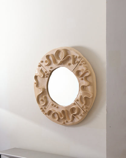 De grand format et de forme circulaire, ce miroir entièrement façonné à la main s’apparente à une pièce de tissu sculptée. Le cadre, divisé en quatre sections visibles, se pare de courbes souples et ondulées, tels des fils qui s’étirent et se croisent à la surface de la matière.
Ces lignes sinueuses évoquent le mouvement d’un ruban, entouré de boutons qui roulent dans les creux du tissu, tandis que la structure suggère les contours d’une crinoline légère.