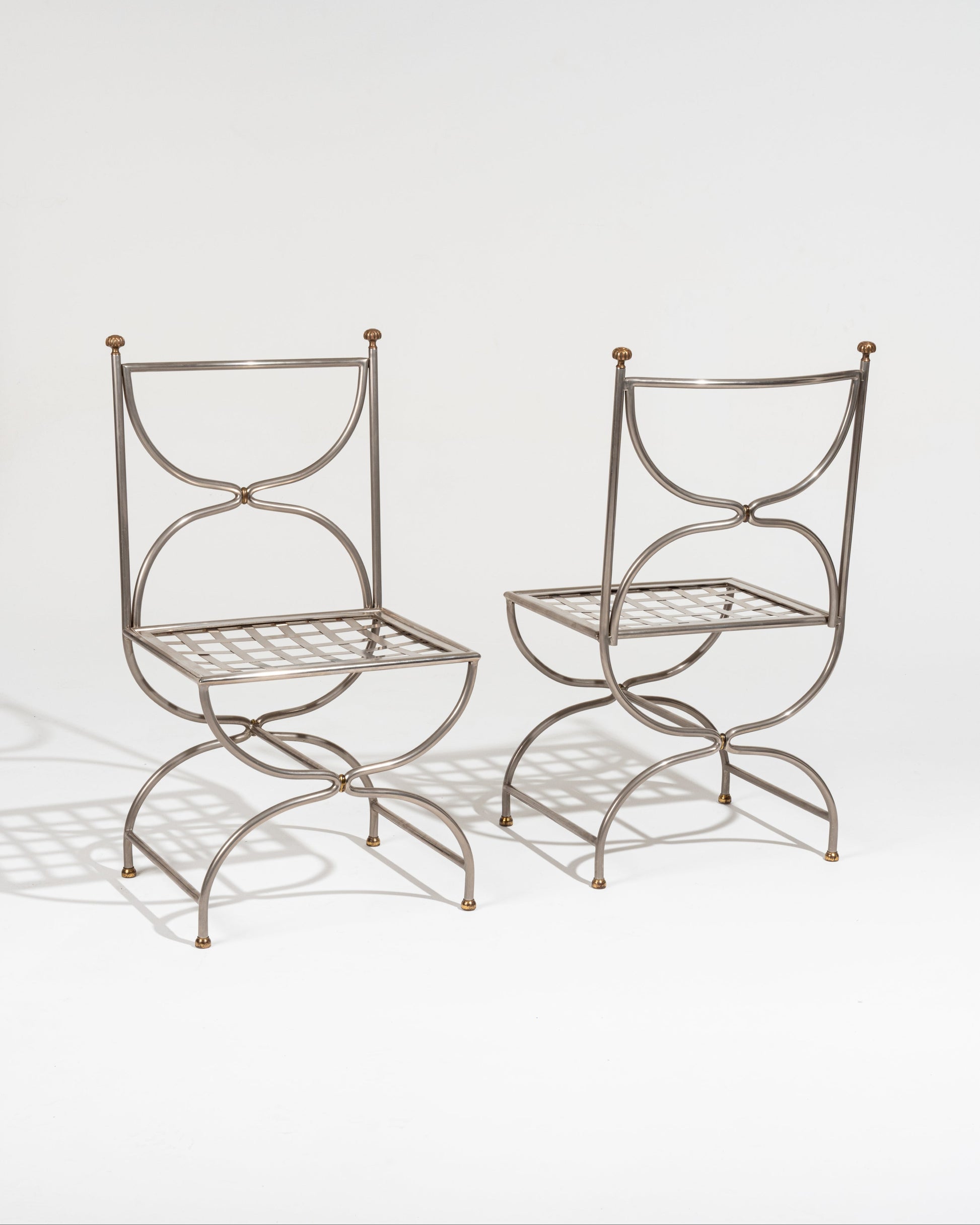 Paire de chaises Maison Jansen en fer forgé datant des années 1980. Le dossier se compose de deux arcs opposés formant une silhouette évoquant un sablier retourné. Les extrémités du dossier sont surmontées de petits pommeaux décoratifs en laiton. L'assise, carrée, est constituée d'un tressage métallique. le piétement se distingue par ses lignes courbes qui se croisent sous l'assise.
Cette pièce est un exemple du travail de la Maison Jansen, mêlant inspirations historiques et modernité décorative. Très bon é