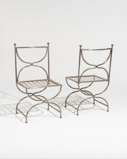 Paire de chaises Maison Jansen en fer forgé datant des années 1980. Le dossier se compose de deux arcs opposés formant une silhouette évoquant un sablier retourné. Les extrémités du dossier sont surmontées de petits pommeaux décoratifs en laiton. L'assise, carrée, est constituée d'un tressage métallique. le piétement se distingue par ses lignes courbes qui se croisent sous l'assise.
Cette pièce est un exemple du travail de la Maison Jansen, mêlant inspirations historiques et modernité décorative. Très bon é