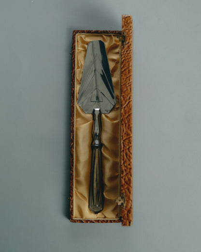 Pelle de service en métal argenté, à décor géométrique typique de la période Art Déco. Manche facetté orné de reliefs symétriques, lame triangulaire finement ciselée. Présentée dans son coffret gainé d'origine. Travail européen, circa 1930-40.

Très bel état général, légère patine conforme à l'âge.