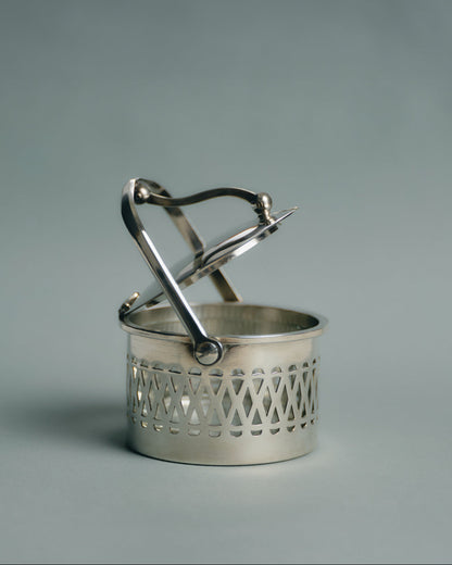 Pot de service à parmesan ou confiture en métal argenté et verre, travail français des années 1950. Monture ajourée à motif de croisillons, couvercle assorti et anse latérale fine. Le récipient intérieur est en verre épais transparent — également appelé verre-confiturier. Anse latérale fine et couvercle assorti.
Très bon état général, légères traces d’usage conformes à l’âge.
