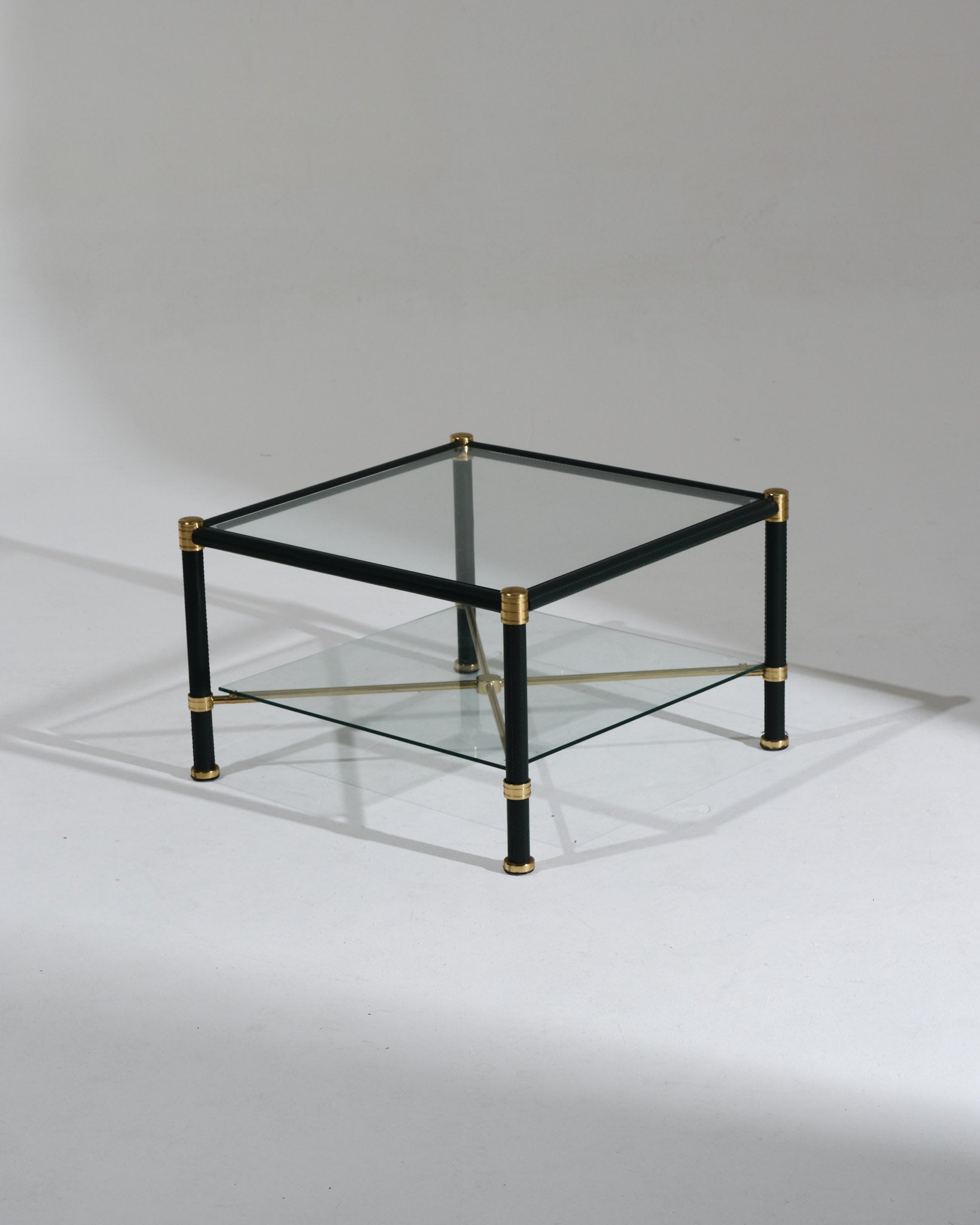 Table basse carrée dans le goût de Jacques Adnet. Travail des années 1950. Structure en laiton gainée de cuir vert foncé à couture sellier, double plateau en verre clair et croisillons centraux en laiton. Parfait état.&nbsp;