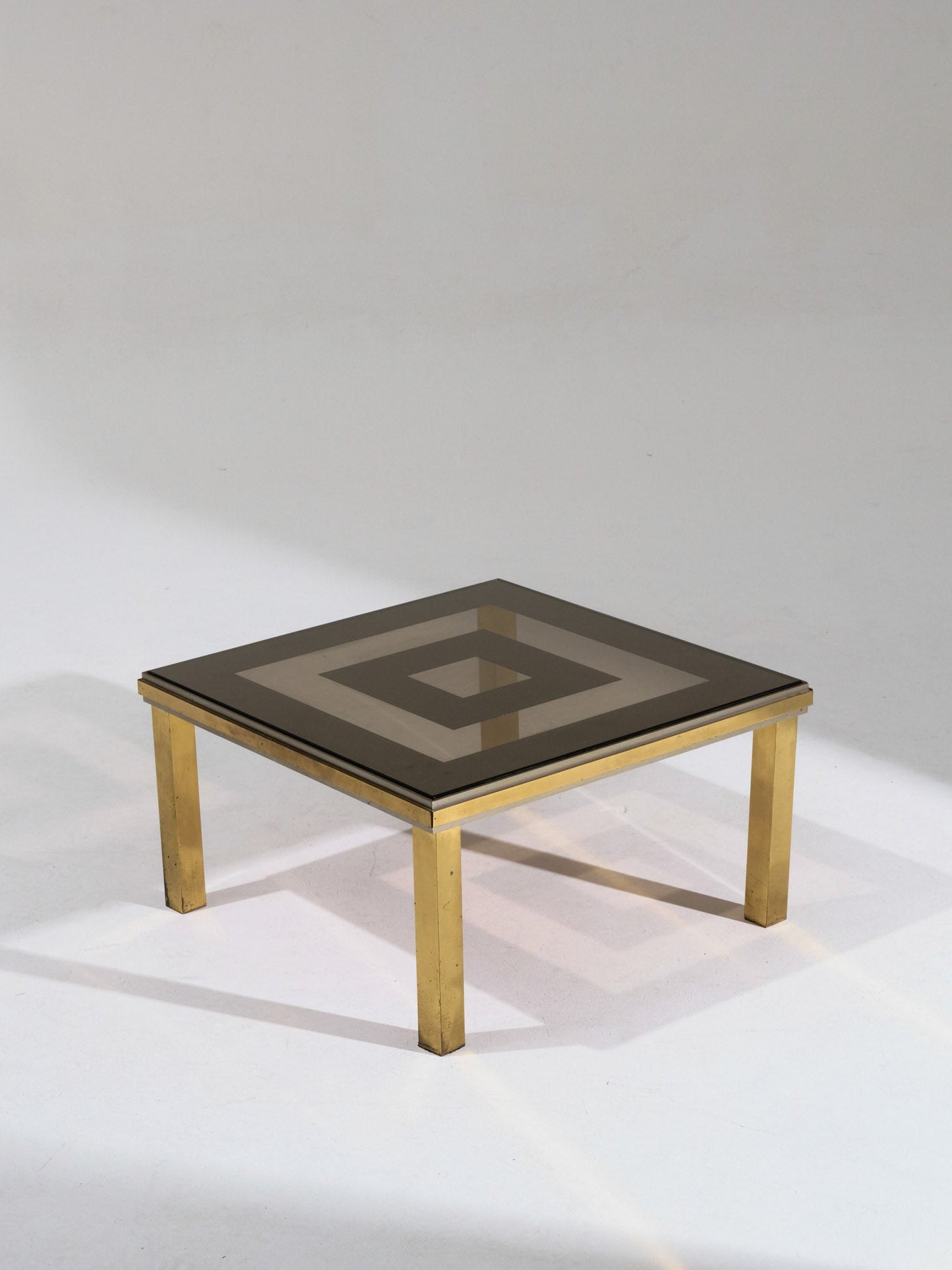Table basse carrée composée d'une structure en laiton poli et d'un double plateau en verre fumé. Les lignes géométriques et la brillance du métal soulignent l'esprit esthétique du design italien des années 1970. Bon état général, légère patine du laiton conforme à l'âge.