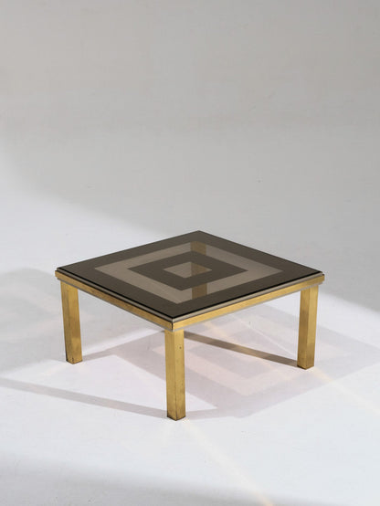 Table basse carrée composée d'une structure en laiton poli et d'un double plateau en verre fumé. Les lignes géométriques et la brillance du métal soulignent l'esprit esthétique du design italien des années 1970. Bon état général, légère patine du laiton conforme à l'âge.