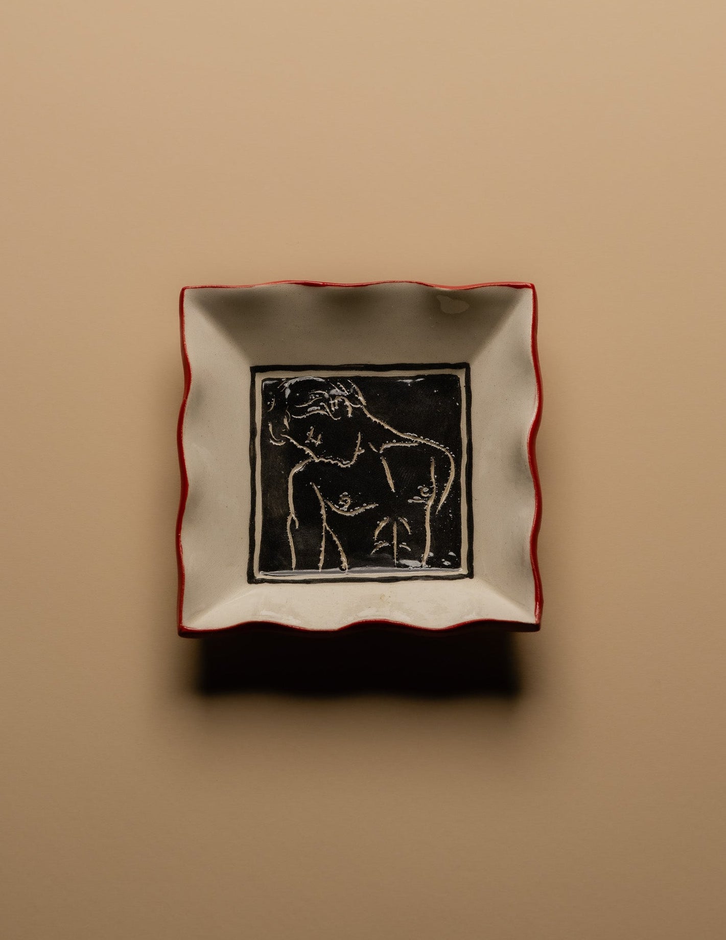 Petit vide-poche carré en céramique émaillée blanche à décor gravé d'un visage masculin entouré de motifs floraux. Pièce unique signée « Eldarov. T » au revers. Contour ondulé souligné d'un liseré rouge. Travail artisanal européen. Parfait état.