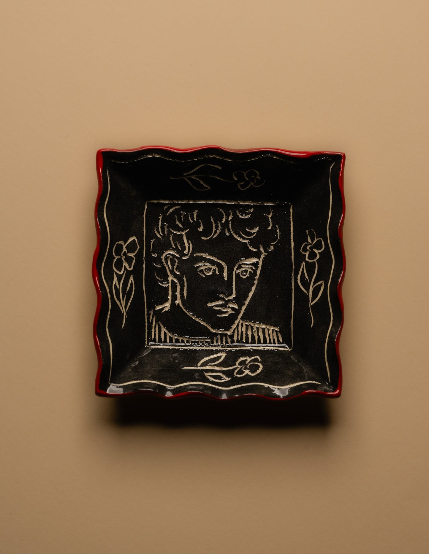 Petit vide-poche carré en céramique émaillée noire à décor gravé d'un visage masculin entouré de motifs floraux. Pièce unique signée « Eldarov. T » au revers. Contour ondulé souligné d'un liseré rouge. Travail artisanal européen. Parfait état.