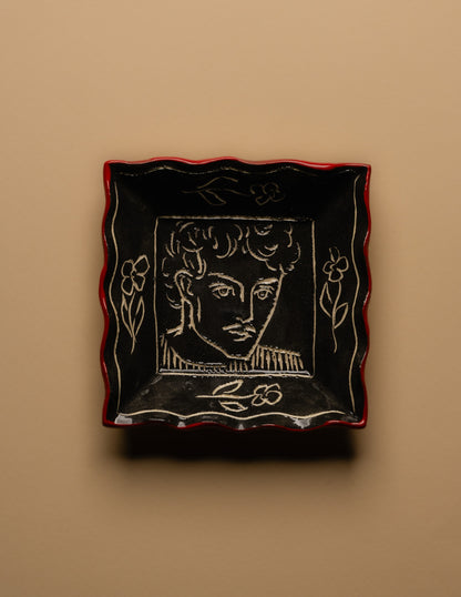 Petit vide-poche carré en céramique émaillée noire à décor gravé d'un visage masculin entouré de motifs floraux. Pièce unique signée « Eldarov. T » au revers. Contour ondulé souligné d'un liseré rouge. Travail artisanal européen. Parfait état.